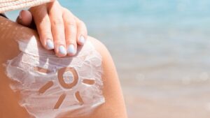 Ruka nanosi kremu za sunčanje na rame, sa nacrtanim suncem u kremi, na plaži pored mora – simbol zaštite kože na suncu